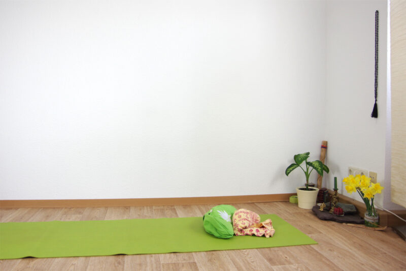 Grüne Yogamatte mit grünem Meditationskissen auf Holzfußboden. Im Hintergrund ein kleiner Altar aus einer Steinplatte mit Kerze, Blumen, Pflanze.